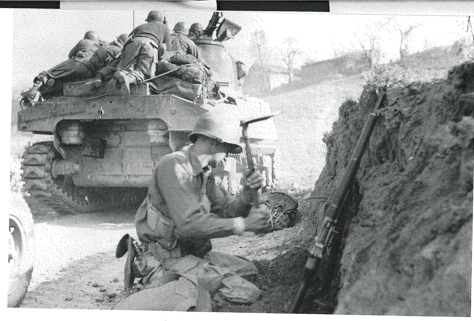 Infantry riding on a tank past a dug-in position in torn-up ground