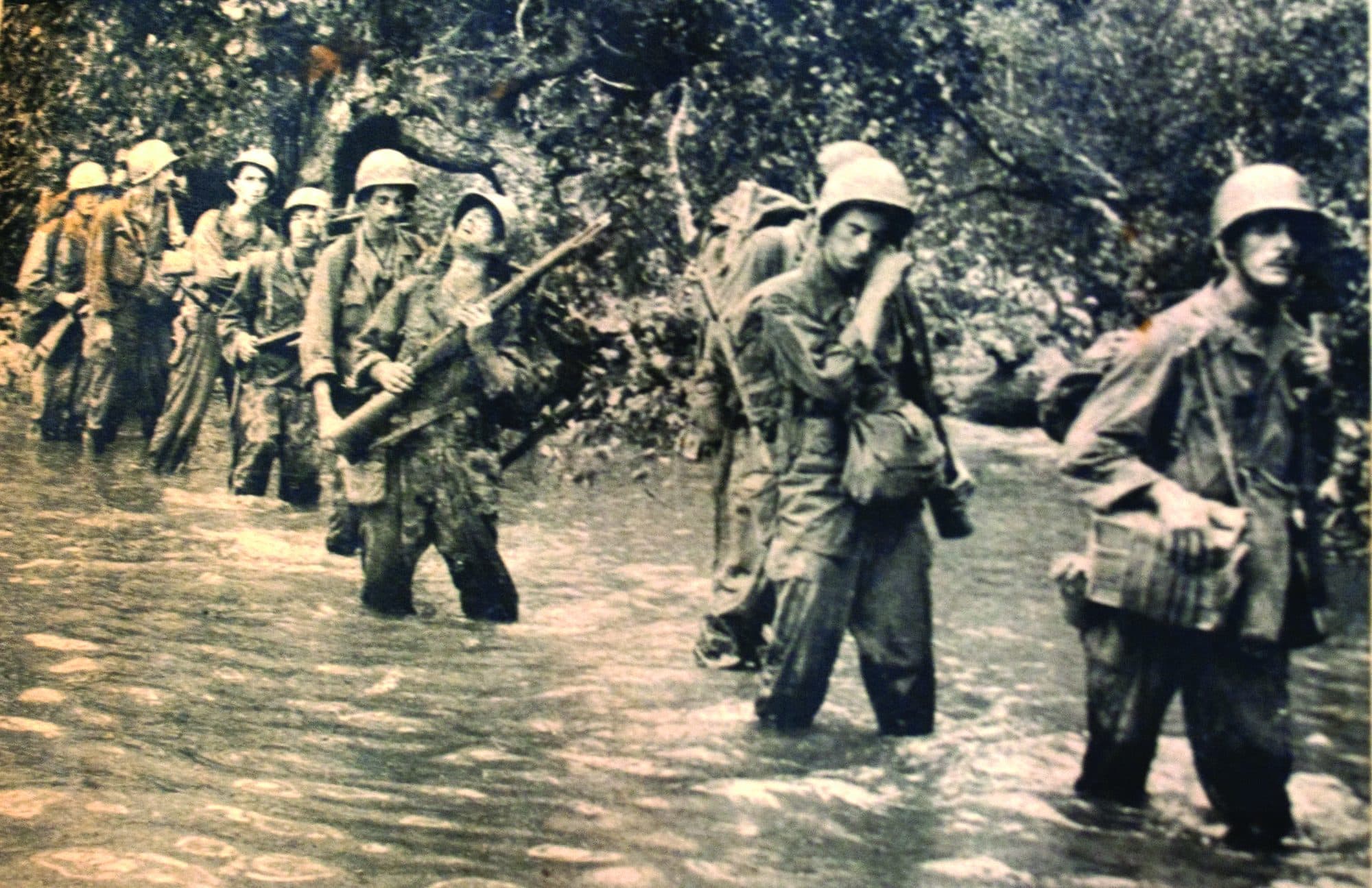 A column of American infantry wading a river through dense jungle, one man looking straight at the camera